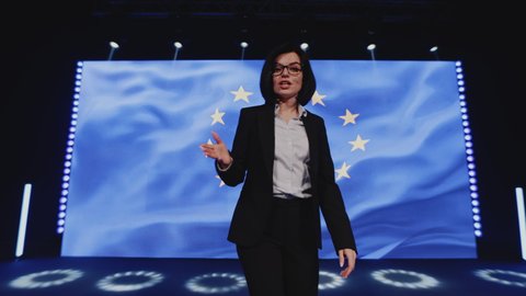 A female TV host talking about the political news of the European Union, the department's conference on climate changes on stage in a studio in front of the european flag on LED screen