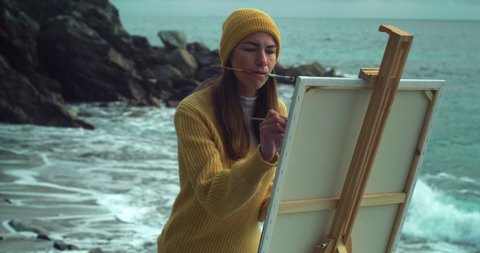 Cinematic shot of young carefree creative female artist painter concentrated and inspired painting with paint brush and oil colors on canvas her new masterpiece on dramatic winter seascape background.