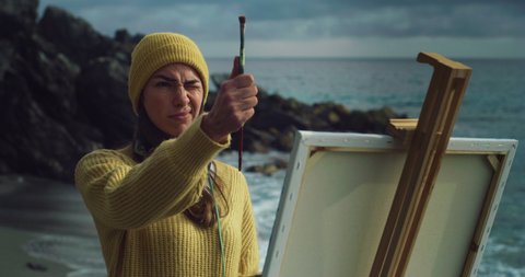 Cinematic shot of young carefree creative female artist painter concentrated and inspired painting with paint brush and oil colors on canvas her new masterpiece on dramatic winter seascape background.