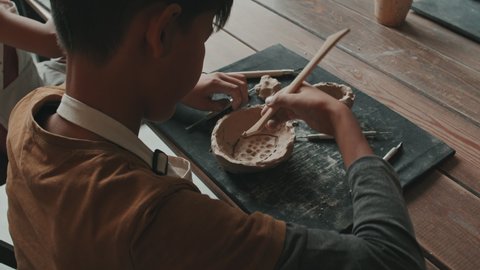 Over-shoulder of little Asian boy sitting at table in pottery school, carving pattern on inside of bowl made of raw clay, learning pottery
