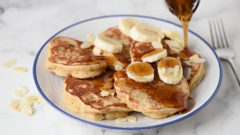 Pouring maple syrup on vegan banana protein oat pancakes. Serving healthy breakfast. Closeup view white marble table