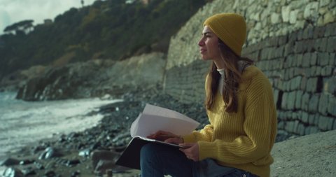 Cinematic shot of young inspired woman is sitting on seashore and writing in pen in notebook romance or letter to friend or lover on dramatic winter seascape with cloudy sky and waves background.