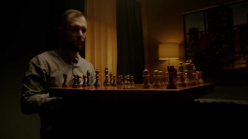 Bearded chessman in shirt moves chess piece pawn sitting at wooden board under electric light in evening slow motion close view
