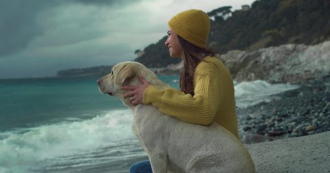 Cinematic shot of young happy smiling woman is caressing with affection her lovely labrador retriever dog puppy while enjoying together dramatic winter seascape together at sunset.
