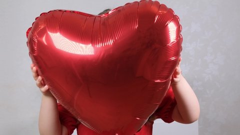 little cute girl in a red dress hides behind a red heart shaped balloon. valentines day concept