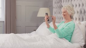 Senior woman at home wearing pyjamas in bed enjoying video call with family on mobile phone - shot in slow motion