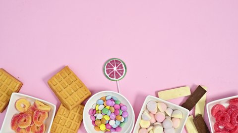 6k Sweet desserts, gummy and chocolate candies and lollypops appear on bottom of pastel pink background. Stop motion animation flat lay