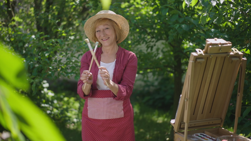 Portrait of confident happy retiree dancing with painting brushes smiling looking at camera. Excited Caucasian woman posing in slow motion having fun enjoying hobby in garden outdoors