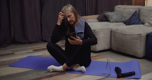Tired Middle Aged Woman after Physical Exercises relaxing on Yoga Mat with Phone. Senior Woman browsing Social Media doing pause before training. Wears black Sportswear. Active Person. Healthy Life.