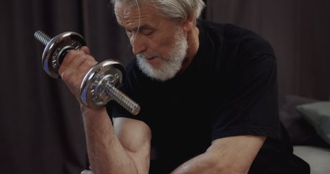 Middle Aged 60s Man holding Dumbbell in hand doing Exercise for Bicep at Home. Having Healthy Habit and Daily Routine. Wears black Sportswear. Strong Fit Older Man. Retirement Activities.