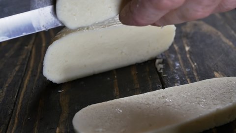 the hands of the cook are cutting soft cheese with a large knife on a wooden brown table for frying in a frying pan. Slow motion, close up