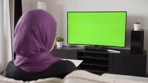 A Muslim woman watches TV with green screen as she sits on a sofa in an apartment - view from behind