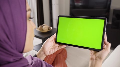 A Muslim woman looks at a tablet (horizontal) with green screen in an apartment - closeup from behind