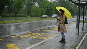 Young beautiful woman using a smartphone and holding a yellow umbrella while waiting for a public transport on a bus stop on a rainy day