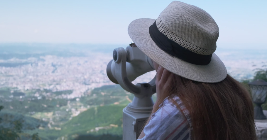 Young woman in hat looks at the city through a telescope, explores sights. Unrecognizable traveler stands on observation deck, examines panorama of city. Travel, adventure opening new horizons. 4k