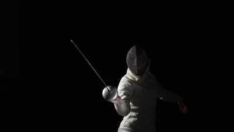 Portrait of young woman fencer in protective helmet becomes fighting position and makes an attacking lunge. An athlete in white uniform poses in dark studio on black background. Slow motion. Close up.