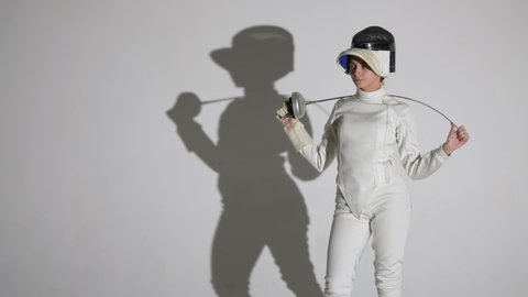 Portrait of a young female fencer removing a protective helmet and holding a sword behind her head. Sportswoman in uniform poses in a dark studio on white background with shadow. Slow motion. Close up