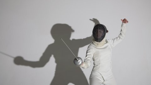 Portrait of a young woman fencer saluting with rapier, putting on helmet and getting into fighting position. An athlete poses in dark studio on white background with shadow. Slow motion.