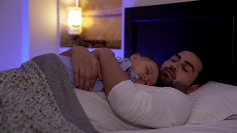 A cute little toddler sleeping peacefully on her father's chest at home. A young Indian dad lovingly kissing his daughter's forehead while resting on the bed - affectionate, relationship and bondin...