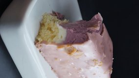 vertical screen. a white porcelain spoon breaks off a purple glazed mousse cake in a white ceramic rectangular bowl