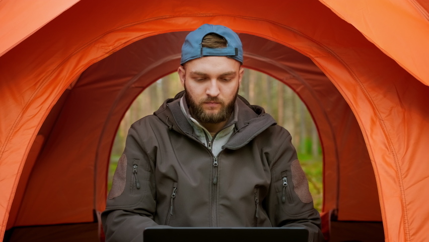 Moving camera back, portrait male freelance tourist remotely working at computer, sitting in an orange tent in nature in woods, drinking hot coffee and working on laptop. Go Everywhere Royalty-Free Stock Footage #1086633506