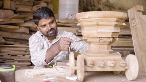 Young indian carpenter polising or shaping chariot by using carpentry tools at workplace - concept of craftperson, self employed and skilled occupation