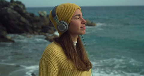 Cinematic shot of young carefree serene woman is putting on headphones to relax her mind by listening her favorite music while breathing fresh air on seashore with dramatic winter seascape at sunset.