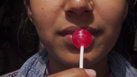 Beautiful young girl eating a red lollipop using his mouth on a dark background. Crop face close-up view.