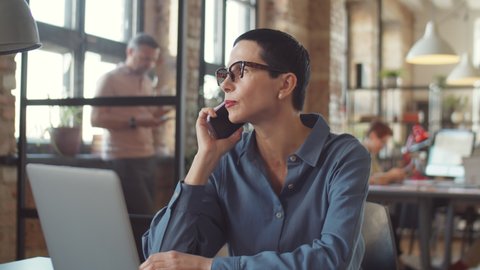 Elegant middle aged businesswoman with short brunette hair talking on mobile phone while sitting at desk in office