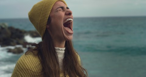 Cinematic close up shot of young desperate tensed furious woman is screaming in loud anger and frustration in the wind to free herself on seashore with dramatic winter seascape at sunset.
