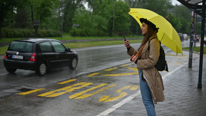 Beautiful woman using a smartphone and holding a yellow umbrella while on the bus stop in the city on a rainy day