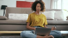 Video of relaxing woman paying something online with her credit card with laptop while sitting on the floor in living room at home.