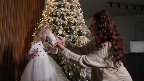 caring parent of female porn gives a drink to little girl after playing and dancing at home backdrop of Christmas tree