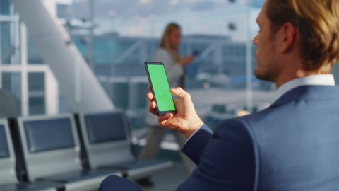 Airport Terminal: Businessman Working on Green Chroma Key Screen Mobile Smartphone, Waits for a Plane Flight. Entrepreneur Does Online Remote Work in Boarding Lounge of Airline Hub. Over the Shoulder