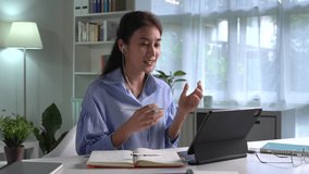 Asian woman wearing earphones having a video call on a tablet sitting on a desk in the living room at home. Young Asian woman entrepreneur giving online educational class lecture, consulting customer