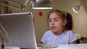 Cute preschool girl is sitting at home at a desk and at a laptop, talking on a video call. Online e-learning class, studying with a teacher, social distance learning, home learning concept.