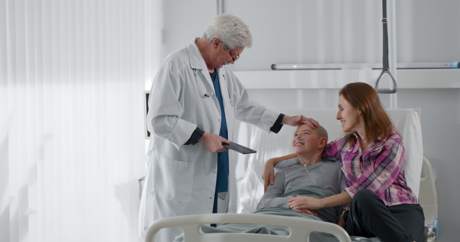 Aged doctor oncologist discharging teen patient with cancer. Happy mother and bald kid talking to doctor and being discharged after chemotherapy sitting in ward | Shutterstock HD Video #1086653951