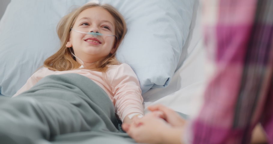 Cute preteen girl lying in hospital bed and holding mother hand. Close up portrait of woman holding hand of sick daughter resting in bed and talking during visit in hospital ward Royalty-Free Stock Footage #1086653987