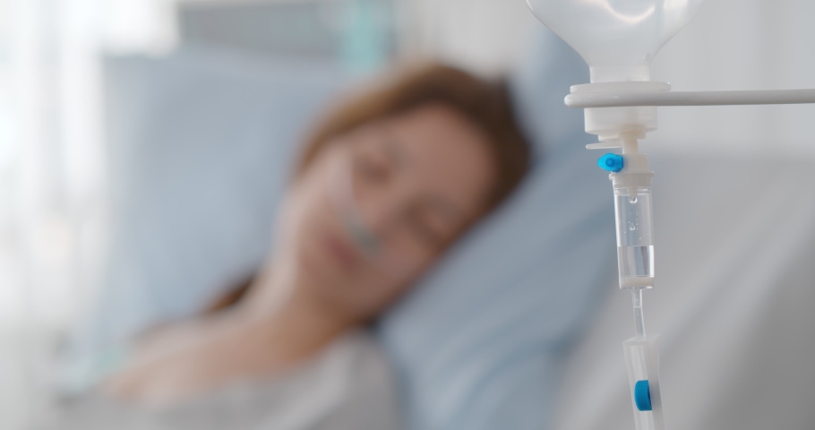 Focus on dripper with medication and sick woman resting in hospital bed on blurred background. Ill female patient feeling unwell sleeping in bed with intravenous dropper in clinic | Shutterstock HD Video #1086653999