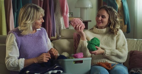 Cinematic close up of mature creative mother and her adult daughter knit together warm yarn wool with knitting needles while sitting comfortably on sofa in living room at home during covid-19 pandemic