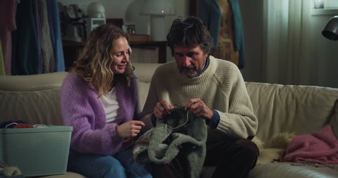Cinematic shot of skilled adult daughter is showing to her mature father how to knit warm yarn wool with knitting needles while sitting comfortably on sofa at home during covid-19 pandemic.