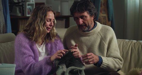 Cinematic close up shot of skilled adult daughter is showing to her mature father how to knit warm yarn wool with knitting needles while sitting comfortably on sofa at home during covid-19 pandemic.