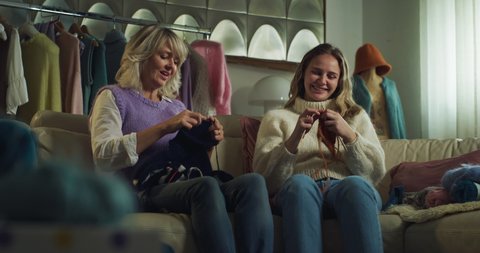 Cinematic shot of mature creative mother is showing design of jumper to her adult daughter for knit together warm yarn wool with knitting needles while sitting comfortably on sofa at home.