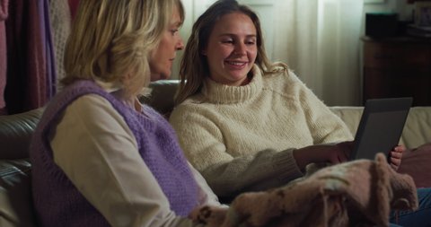 Cinematic shot of mature creative mother and adult daughter choosing together on tablet design of warm clothes to be knitted with yarn wool on sofa at home during covid-19 pandemic lockdown.
