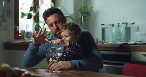 Cinematic shot of young father is showing to his curious happy little toddler boy how to make music sounds with filled crystal glasses of water with fingers while playing together in kitchen at home.