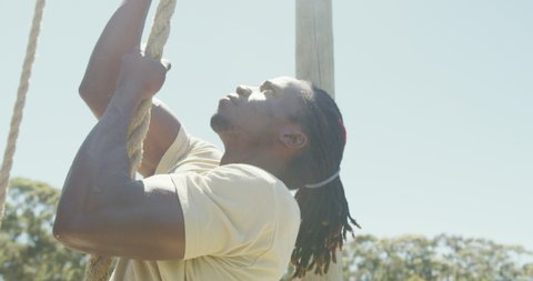 Fit african american male soldier with deadlocks climbing rope on obstacle course in sun. healthy active lifestyle, cross training outdoors at boot camp.