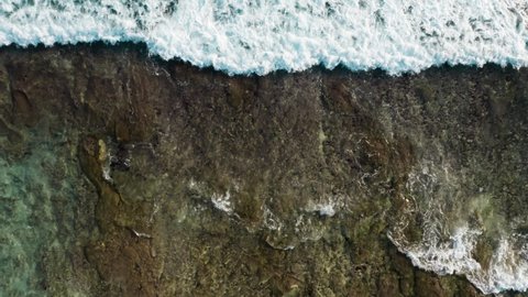 Static aerial footage of brown reef with azure water covered by waves crashing into white sea foam water. Copter footage of tidal surf washing rocky ocean shore. Drone shot of stone and water texture