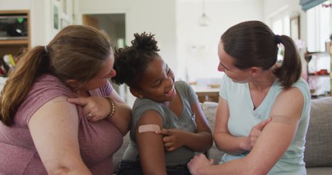 Caucasian lesbian couple and their african american daughter pointing at bandages on their arms. domestic life, spending time at home during covid 19 pandemic.