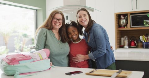 Portrait of happy caucasian lesbian couple and their african american daughter embracing in kitchen. domestic life, spending time at home.