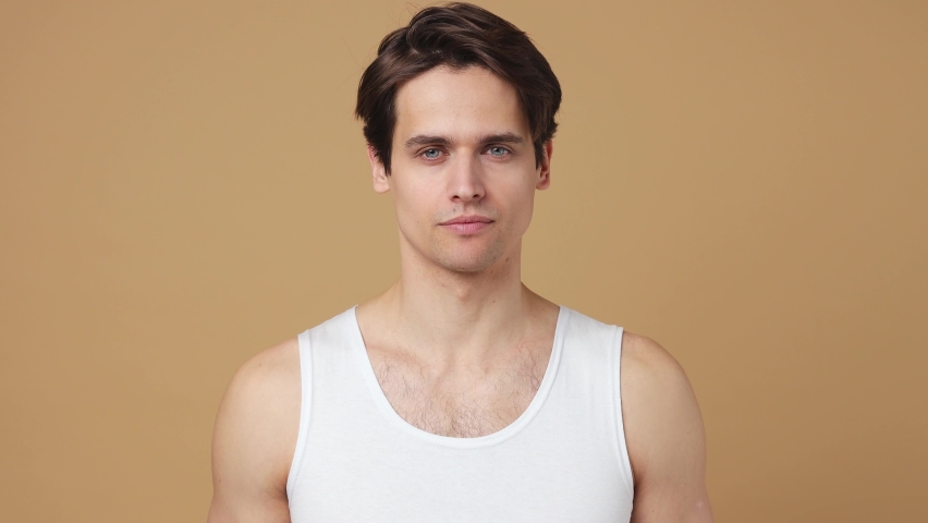 Happy young man 20s perfect skin wears white tank top look camera like in mirror brush teeth isolated on plain pastel beige background studio portrait. Body care healthcare cosmetic procedures concept Royalty-Free Stock Footage #1086675836